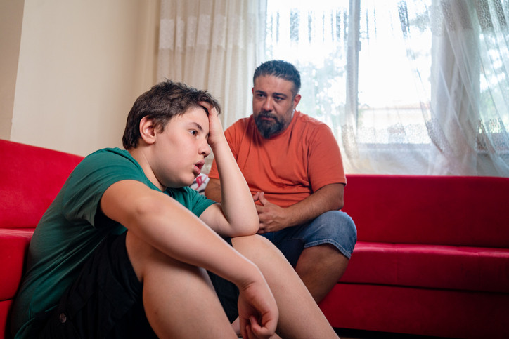 A young boy sitting by a red couch with his head resting on his hand, looking distressed, while an adult man sits nearby co-regulating him.